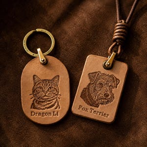 Könnte beinhalten: Ein ovaler Schlüsselanhänger aus beigem Leder mit eingeprägter Dragon Li Katze und ein rechteckiger Lederanhänger an einer braunen Kordel mit eingeprägtem Fox Terrier Hund. Beide Artikel haben goldfarbene Metallbeschläge und Gravuren.