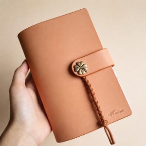 May include: A tan leather notebook cover featuring a smooth finish and a strap closure secured by a brass-toned four-leaf clover button. A braided leather cord hangs from the closure, and the name Karen is embossed in the bottom right corner.