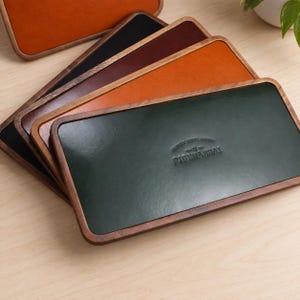 May include: A stack of five rectangular valet trays with wooden frames and leather inserts in dark green, orange, dark brown, black, and light orange. The top dark green tray features the embossed text PIGENANHAY in the center of the leather surface.