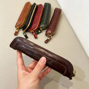 Può includere: Una collezione di astucci in pelle per penne in vari colori, tra cui marrone, nero, rosso e verde. Ogni astuccio presenta una cerniera dorata e un piccolo tirante in pelle. Un astuccio è tenuto in mano, mostrando le cuciture dettagliate e la consistenza della pelle.