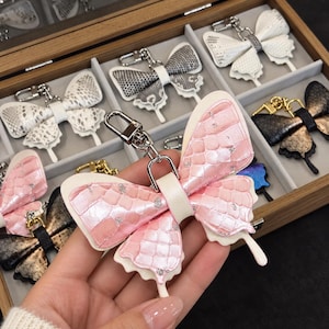 May include: A pink butterfly-shaped keyring with a textured, iridescent scale pattern and silver glitter accents. The keyring features a silver metal lobster clasp and is displayed alongside other butterfly keyrings in a wooden box.