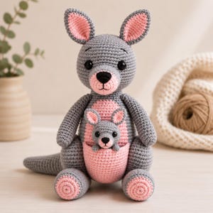 Op de afbeelding: Een gehaakte kangoeroe knuffel in grijs en lichtroze, bestaande uit een grote volwassen kangoeroe met een buidel waarin een kleinere babykangoeroe zit. Beide figuren hebben zwarte veiligheidsoogjes, zwarte neuzen en roze details.