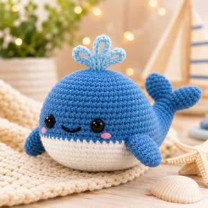 May include: A crocheted blue whale with a white belly, small fins, and a tail. The whale has black eyes, pink cheeks, and a small spout on top. It is sitting on a cream-colored crocheted blanket.