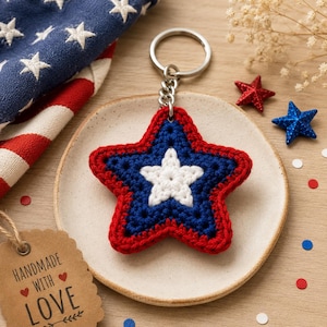 May include: A crocheted star keychain featuring a white center star on a blue background with a red border, attached to a silver metal ring. The design includes a brown tag with the text "HANDMADE WITH LOVE" and small red heart icons.