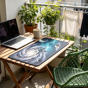Può includere: Un laptop è appoggiato su un tavolo di legno con un tappetino per mouse a tema galassia. Il tappetino presenta un design a galassia a spirale con pianeti e un emblema a forma di bussola. Una sedia verde e piante in vaso sono sullo sfondo.