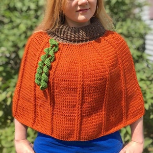 Puede incluir: Un poncho de punto de ganchillo con forma de calabaza, con un cuello alto marrón y un tallo de ganchillo verde. El poncho es naranja y lo lleva una persona con el pelo largo y rubio.