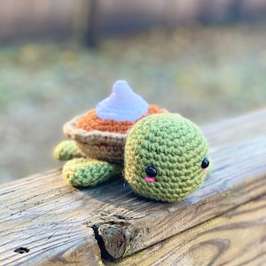 以下が含まれることがあります： 茶色とオレンジの殻と白い編み物でできたトッピングが付いた、かぎ針編みのかめ。かめは緑色で、黒い目にピンクの頬をしています。かめは木製の表面に座っています。