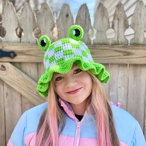 May include: A green and white crocheted bucket hat with frog eyes and a ruffled brim. The hat is worn by a person with pink hair and a multi-colored jacket.