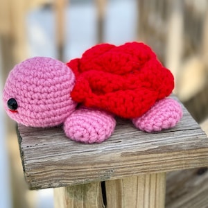 Könnte beinhalten: Eine gehäkelte rosa und rote Schildkröte mit schwarzen Augen. Die Schildkröte sitzt auf einer Holzoberfläche.