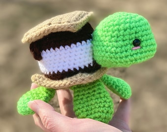 Puede incluir: Una tortuga de crochet verde con un s'more de crochet marrón, blanco y beige en su espalda.