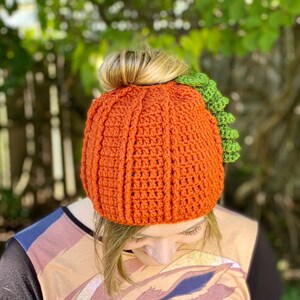 May include: A crocheted pumpkin hat with a green stem. The hat is orange and has a textured design. The hat is worn by a person with brown hair.