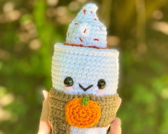 May include: A crocheted white coffee cup with a brown scarf and a crocheted orange pumpkin. The cup has a cute face with black eyes and a smile.