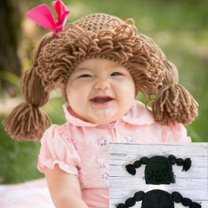 May include: A baby wearing a brown crocheted hat with pigtails. The hat is shaped like a doll's head with a pink bow on top. The baby is smiling and looking at the camera.