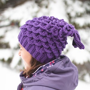 May include: A purple crocheted hat with a dragon scale pattern. The hat has a long tail that hangs down the back of the neck. The hat is worn by a person in a purple jacket. The text "Sweet Moments by Wendy" is visible at the bottom of the image.