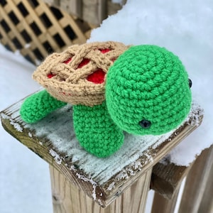 以下が含まれることがあります： 緑色の甲羅と茶色のパイ生地の甲羅を持つ、かぎ針編みで作った亀のぬいぐるみ。亀は雪で覆われた木の柵の支柱の上に座っている。