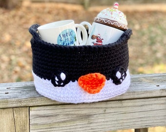 May include: A crocheted penguin-shaped basket with a white rim and orange beak. The basket is filled with a white mug with the text "Snow much fun" and a white ceramic jar with a white and red frosting-like topping.
