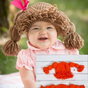 May include: A baby wearing a brown crocheted doll hat with pigtails. The hat has a pink bow on top. Two orange crocheted doll hats with pigtails are shown on a white wooden surface.