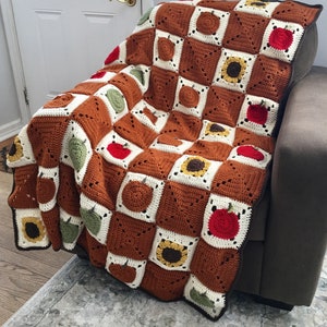 May include: A crocheted throw blanket with a checkered pattern featuring sunflowers, apples, and pumpkins in shades of brown, green, red, and white.