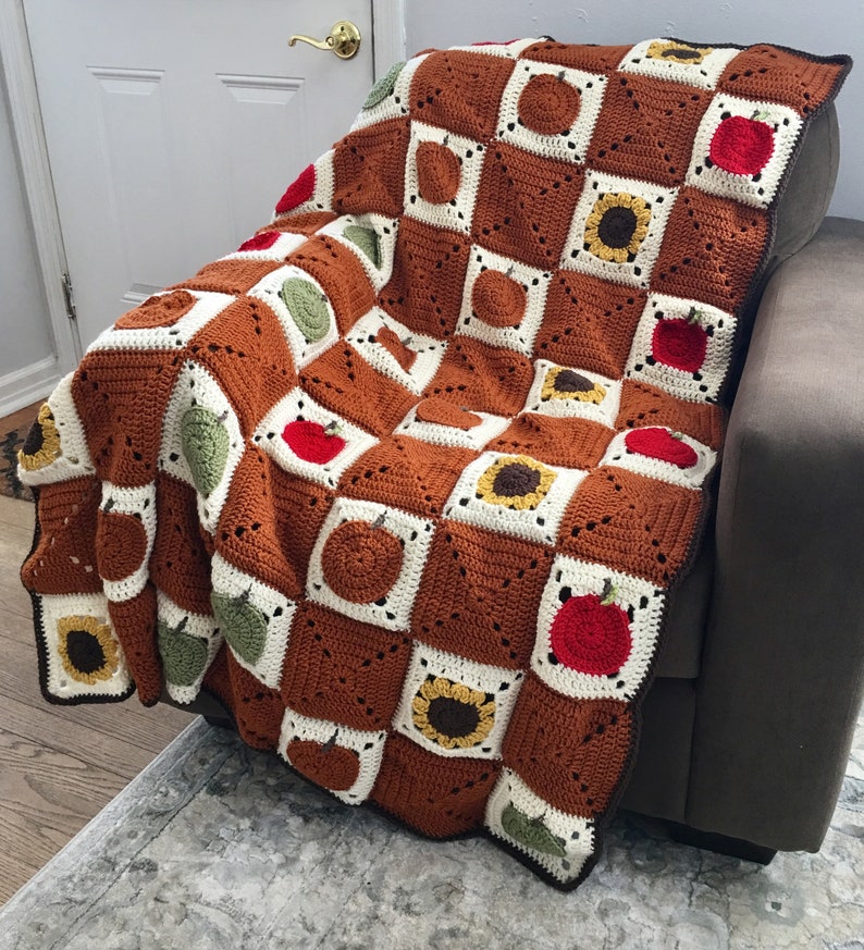 May include: A crocheted throw blanket with a checkered pattern featuring sunflowers, apples, and pumpkins in shades of brown, green, red, and white.