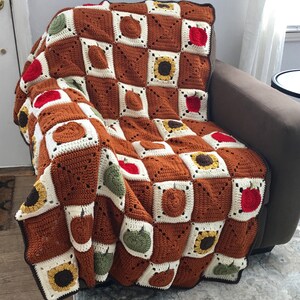 May include: A crocheted blanket with a brown, white, green, yellow, and red color scheme. The blanket features a repeating pattern of pumpkins, apples, and sunflowers.