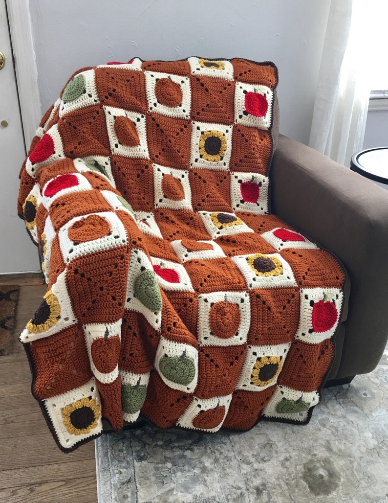 May include: A crocheted blanket with a brown, white, green, yellow, and red color scheme. The blanket features a repeating pattern of pumpkins, apples, and sunflowers.