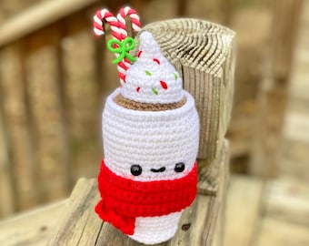 May include: A crocheted plush toy shaped like a cup of hot chocolate with a red scarf, white whipped cream, and candy cane stirrers.