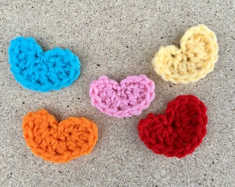 May include: Five crocheted hearts in different colors: blue, pink, yellow, orange, and red. The hearts are arranged in a semi-circle on a brown textured surface.