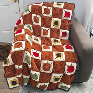 May include: A crocheted throw blanket with a checkered pattern featuring sunflowers, apples, and pumpkins in shades of brown, yellow, green, and red.