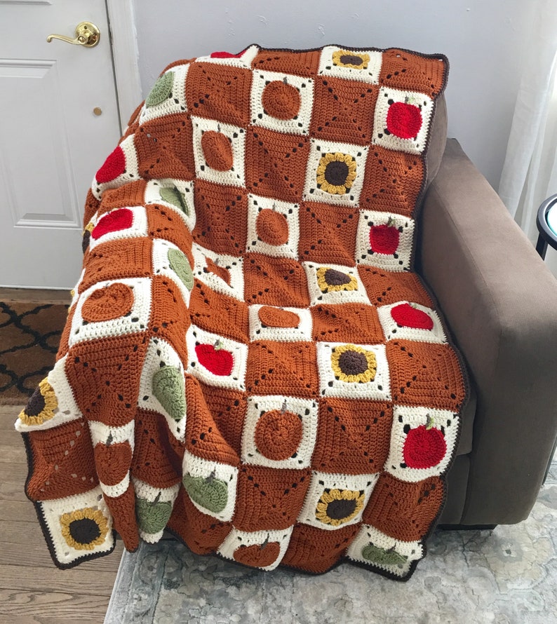 May include: A crocheted throw blanket with a checkered pattern featuring sunflowers, apples, and pumpkins in shades of brown, yellow, green, and red.