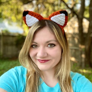 May include: A woman wearing a crocheted fox headband with orange and white fur. The headband has black eyes and a white nose.