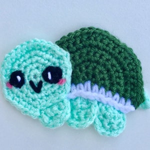 Könnte beinhalten: Eine gehäkelte Schildkröte mit grünem Panzer und hellblauem Körper. Die Schildkröte hat schwarze Augen und einen rosa Rouge auf den Wangen.