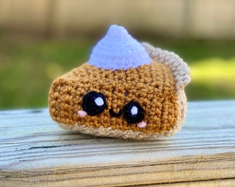 May include: A crocheted pumpkin pie with a purple whipped cream topping and a cute cartoon face. The pie is brown and has a white crust.