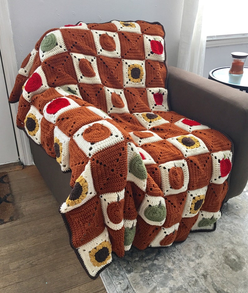 May include: A crocheted throw blanket with a checkered pattern featuring apples, sunflowers, and pears in shades of brown, red, green, and yellow.