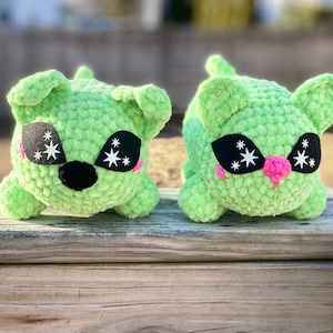May include: Two green crocheted plush toys with black eyes and white stars. The toys have pink noses and are sitting on a wooden surface.