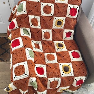 May include: A crocheted throw blanket with a checkered pattern featuring apples, pumpkins, and sunflowers. The blanket is brown, white, green, yellow, and orange.