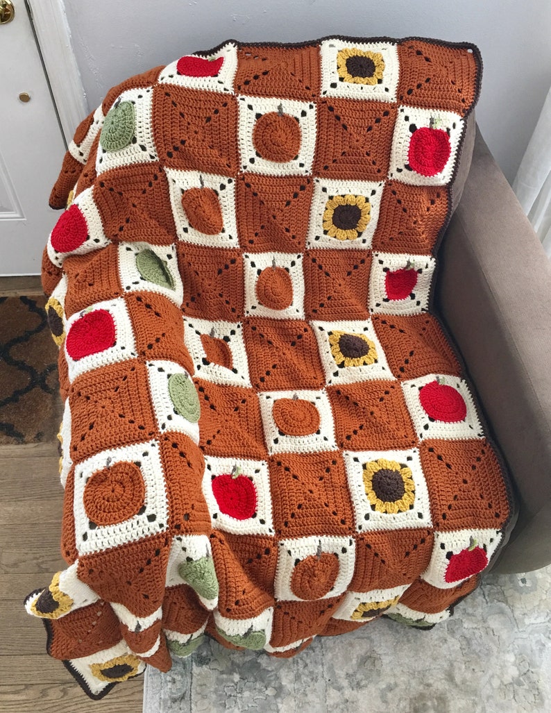 May include: A crocheted throw blanket with a checkered pattern featuring apples, pumpkins, and sunflowers. The blanket is brown, white, green, yellow, and orange.
