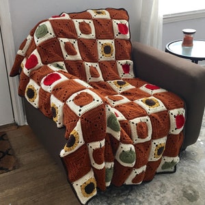 May include: A crocheted throw blanket with a checkered pattern in brown, white, green, and red. The blanket features a sunflower design in each square.