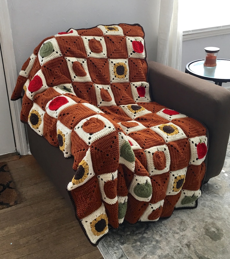 May include: A crocheted throw blanket with a checkered pattern in brown, white, green, and red. The blanket features a sunflower design in each square.