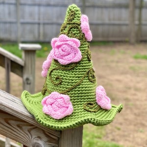 Könnte beinhalten: Ein grüner Häkelhut mit rosa gehäkelten Rosen und grünen Ranken. Der Hut hat eine spitze Krone und eine breite Krempe.