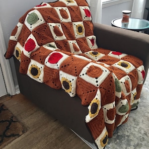 May include: A brown, white, and green crocheted throw blanket with a sunflower pattern. The blanket is draped over a brown armchair.