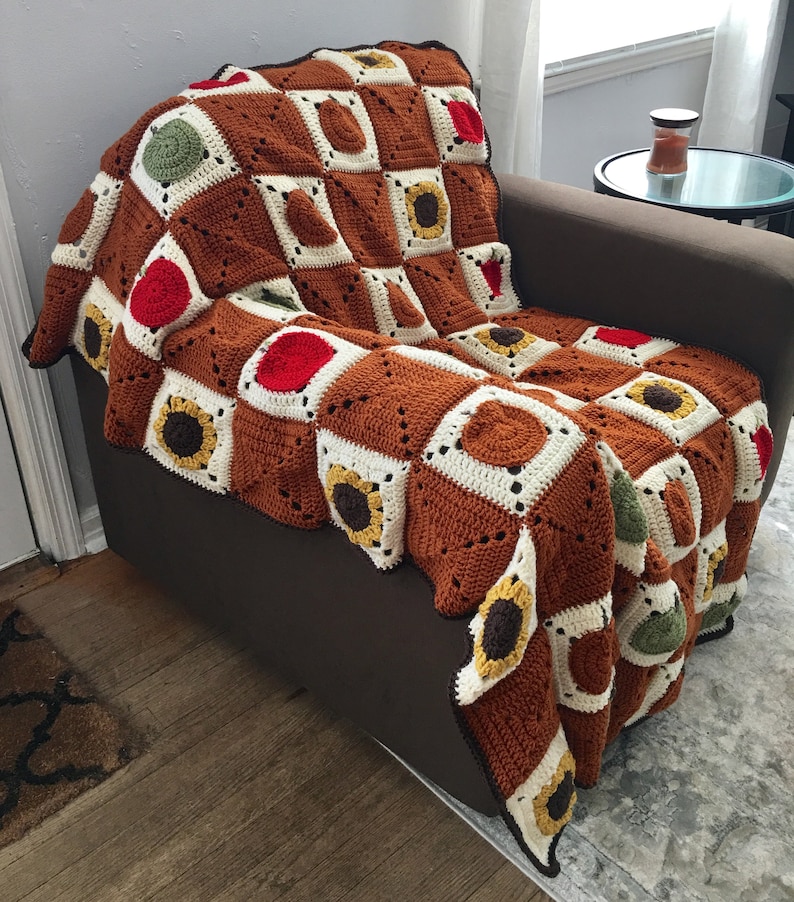 May include: A brown, white, and green crocheted throw blanket with a sunflower pattern. The blanket is draped over a brown armchair.