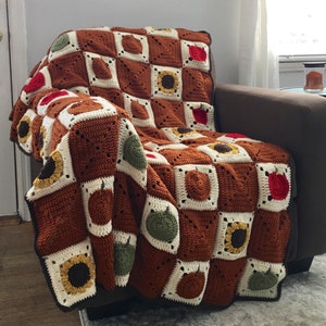 May include: A crocheted throw blanket with a brown, white, and green checkered pattern. The blanket features various motifs, including sunflowers, apples, and pears.