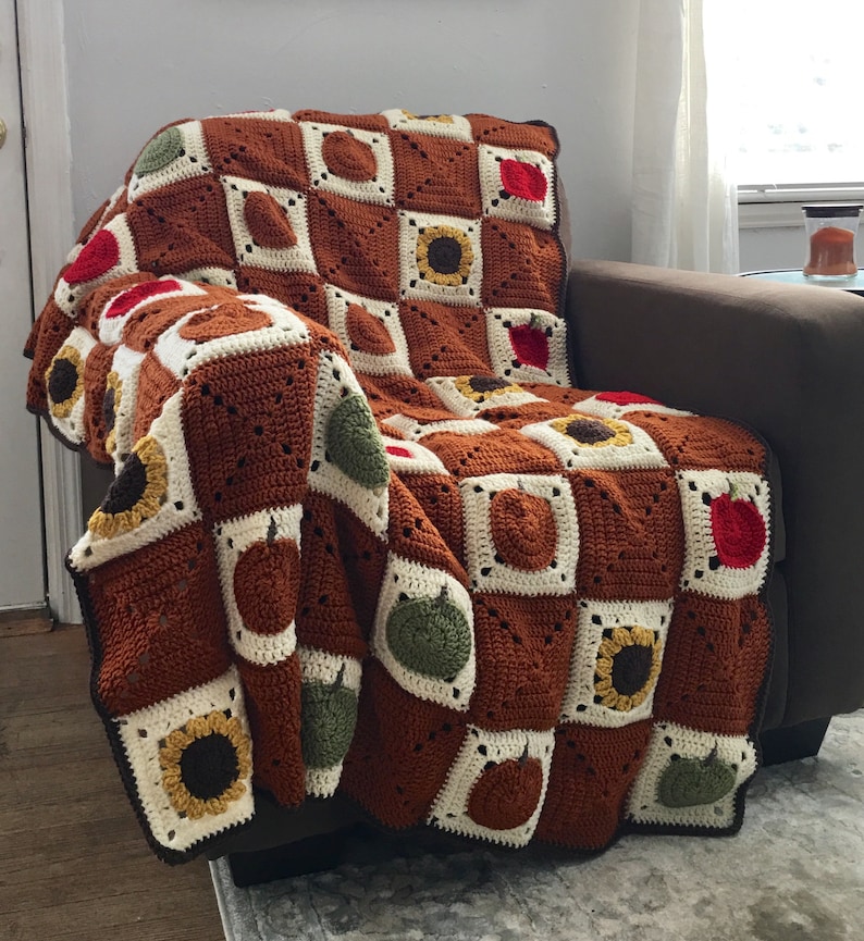May include: A crocheted throw blanket with a brown, white, and green checkered pattern. The blanket features various motifs, including sunflowers, apples, and pears.