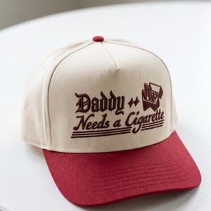 May include: A beige and burgundy baseball cap with the text "Daddy Needs a Cigarette" in brown. The cap features a burgundy brim and a small burgundy button on top. The design includes an illustration of a pack of cigarettes.