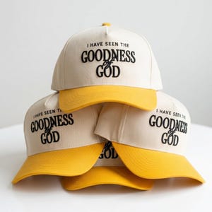 May include: A stack of beige baseball caps with yellow brims. Each cap has the text "I HAVE SEEN THE GOODNESS OF GOD" in black lettering. The caps are stacked on top of each other.