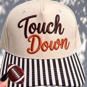 May include: A cream-colored baseball cap featuring the embroidered text Touch Down in dark brown and orange script. The brim displays a vertical black and white stripe pattern, and a small brown football patch is stitched onto the left side.