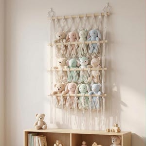 Op de afbeelding: Een macramé wandplank toont knuffels, waaronder teddyberen en konijntjes, in pastelkleuren. De plank heeft drie houten niveaus en is opgehangen aan een geweven touwontwerp. Een kleine teddybeer zit op een houten plank hieronder.