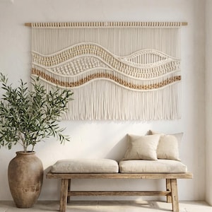 Op de afbeelding: Een grote, crèmekleurige macramé wandhanger met een golvend ontwerp, geaccentueerd met bruine en gouden kralen. Daaronder een houten bank met beige kussens en kussens. Een grote, bruine vaas bevat een groene plant.