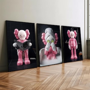 May include: Three framed art prints featuring pink cartoon figures in bathroom settings. One figure reads a newspaper on a toilet, another relaxes in a bathtub with cucumber slices on its eyes, and the third stands in a shower.