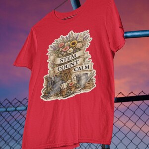 Peut inclure: Un t-shirt rouge à manches courtes avec une impression graphique représentant un bouquet de fleurs, des outils de jardinage et un arrosoir. Une bannière en ruban beige affiche le texte STEM COUNT CALM en lettres capitales noires.
